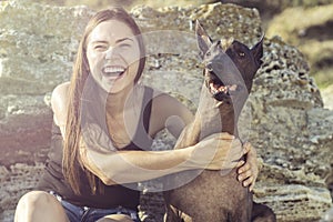 Cheerful pretty young girl sitting and hugging her dog xoloitzcuintli on the stoun beach at sunset