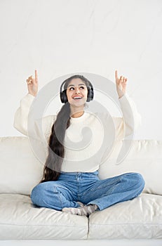 Cheerful pretty young woman in headphones showing her fingers up on empty wall while sitting on couch