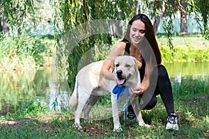 Cheerful pretty young woman sitting and hugging her dog at river bank