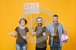 Cheerful protesting young three people hold protest broadsheet placard plastic bottles trash bag isolated on yellow photo