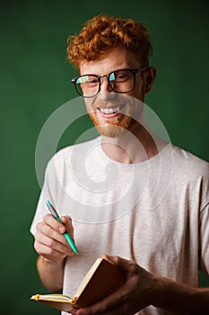 Cheerful readhead bearded student in white tshirt, holding notebook with pen, looking at camera