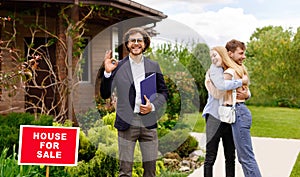 Cheerful realtor showing OK gesture and young happy couple hugging near their new house