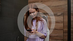 Cheerful red-haired young woman enjoying using mobile phone sitting on toilet at home bathroom with modern interior.