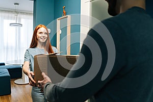 Cheerful red-haired young woman receiving cardboard box from unrecognizable delivery man on doorstep at apartment.