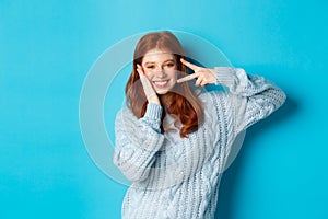 Cheerful redhead female model sending good vibes, smiling and showing peace sign, standing over blue background