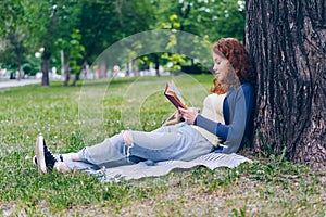 Cheerful redhead girl reading book outdoors in park smiling relaxing on grass