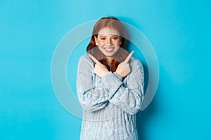 Cheerful redhead girl showing two choices, pointing sideways and smiling over blue background