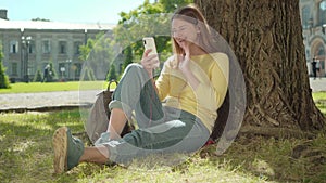 Cheerful redhead girl using video chat outdoors on sunny summer day. Portrait of positive young Caucasian woman sitting