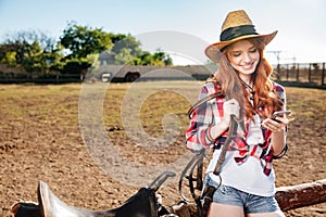 Cheerful redhead young woman cowgirl using cell phone in village