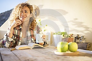 Cheerful relaxed beautiful curly blonde caucasianwoman enjoy the sunny day at home on the terrace reading a book and drinking tea