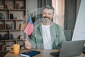 Cheerful retired european man with beard teacher hold USA flag shooting video lesson on laptop