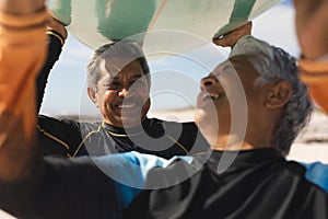 Cheerful retired multiracial senior couple enjoying while carrying surfboard over heads at beach