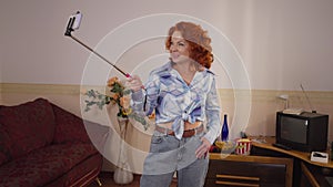 Cheerful retro woman photographing on smartphone with selfie stick in 1990s. Happy confident Caucasian redhead lady