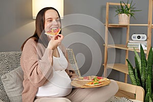 Cheerful satisfied pregnant woman biting big slice of pizza enjoying unhealthy nutrition relaxing at home with delicious fast food