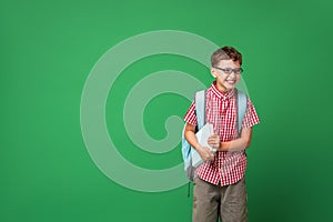 Cheerful schoolboy with glasses, holding book and backpack on green background