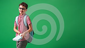 Cheerful schoolboy with glasses, holding book and backpack on green background