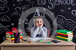 Cheerful schoolboy sitting on the desk with books, school supplies, with both arms under the cheecks