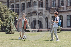 Cheerful schoolboys with backpacks playing football on lawn near school photo