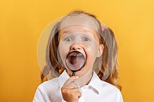 Cheerful schoolgirl little girl shows teeth and smiles through a magnifying glass.