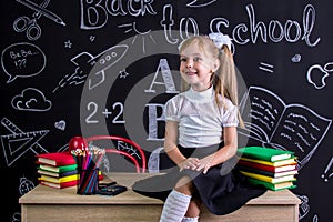 Cheerful schoolgirl sitting on the desk with books, school supplies