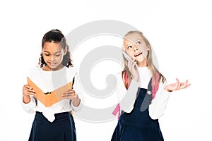 cheerful schoolgirl talking on smartphone near african american friend reading book isolated on white.