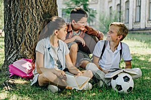 Cheerful schoolkids talking while sitting on lawn under tree