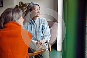 Cheerful senior Asian woman with friend look out of window at table in cozy cafe