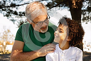 Cheerful senior caucasian grandfather hugging little mixed race boy, talking, enjoy free time, fun outdoor, close up