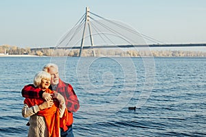 Cheerful senior citizens woman and man are standing and hugging on the lake, against the background of the bridge
