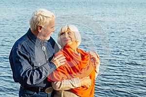 Cheerful senior citizens woman and man are standing and hugging on the lake, against the background of the bridge