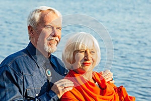 Cheerful senior citizens woman and man are standing and hugging on the lake, against the background of the bridge