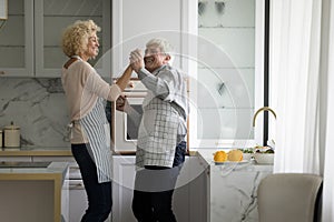 Cheerful senior couple in aprons dancing to music at kitchen