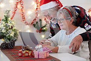 Cheerful senior couple embracing in Christmas sweeter and santa hat while using laptop computer. Elderly happy couple celebrate
