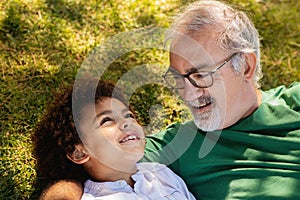 Cheerful senior man with beard and black little boy hugging, have fun, relaxing in park, lying on grass