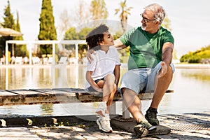 Cheerful senior man with beard and black little boy sitting on bench, relaxing in park, talk