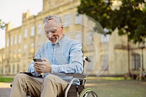 Cheerful senior man, recovering male patient in wheelchair using smartphone while resting alone in the park near
