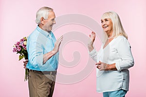 cheerful senior man waving hand while looking at wife and holding bouquet on pink.
