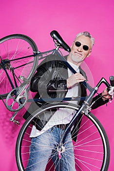 Cheerful senior man wearing leather jacket and sunglesses holding bicycle on his shoulders