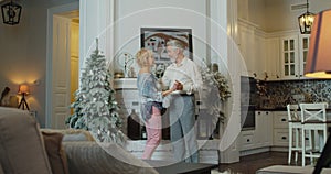 Cheerful senior woman dancing with husband in front of decorated Christmas tree and imitating singing. Funny retirees