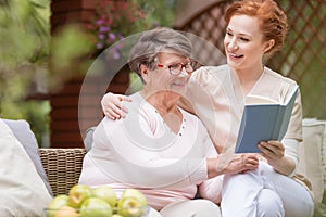 Cheerful senior woman with her tender caretaker reading a book t