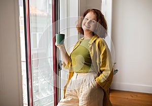 Cheerful senior woman holding green mug standing by window at home