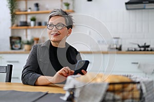 Cheerful senior woman with a mobile phone in her hands in the kitchen smiling at the dining table