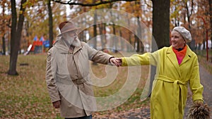 Cheerful senior woman pulling hand of man walking along autumn park alley smiling talking. Positive happy Caucasian old