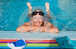 Cheerful senior woman with swimming goggles enjoys water of the swimming pool.  Outdoor pool with clear water. Feet out of water.