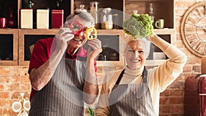 Cheerful seniors fooling with fresh vegetables at kitchen