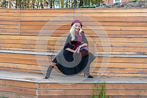 Cheerful sits on a summer theater bench, wooden in a burgundy coat and biret, with a charismatic appearance, in the fall