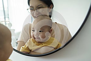 Cheerful smiling Asian mother and little newborn baby son having fun playing and looking at their faces in mirror at home