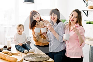 Cheerful smiling attractive young women, middle aged woman and little cute baby girl eating cookies and cupcakes and