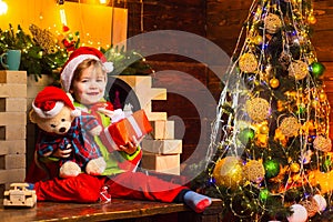 Cheerful smiling baby Santa boy holding his teddy bear and Christmas present near fire place and decorated New Year tree