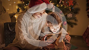 Cheerful smiling boy with mother unwrapping and looking inside box with Christmas presents and gifts. Families and